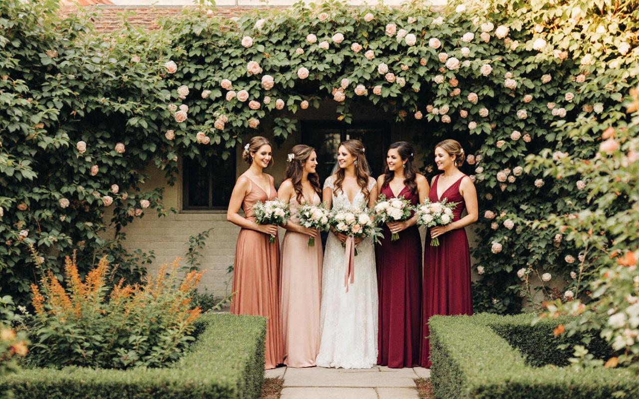 Quatre demoiselles d'honneur en robes cuivre et bordeaux, coiffures soignées et accessoires élégants, tenant de petits bouquets, posant dans un jardin verdoyant baigné d'une douce lumière dorée.