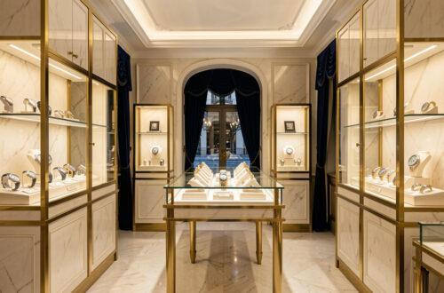 Intérieur élégant d’une bijouterie horlogère parisienne, avec des vitrines lumineuses présentant des montres raffinées et des détails en laiton sur un décor de marbre clair et de velours bleu nuit.