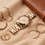 Collection élégante de bijoux en or, montre raffinée et articles de maroquinerie disposés sur un velours beige.